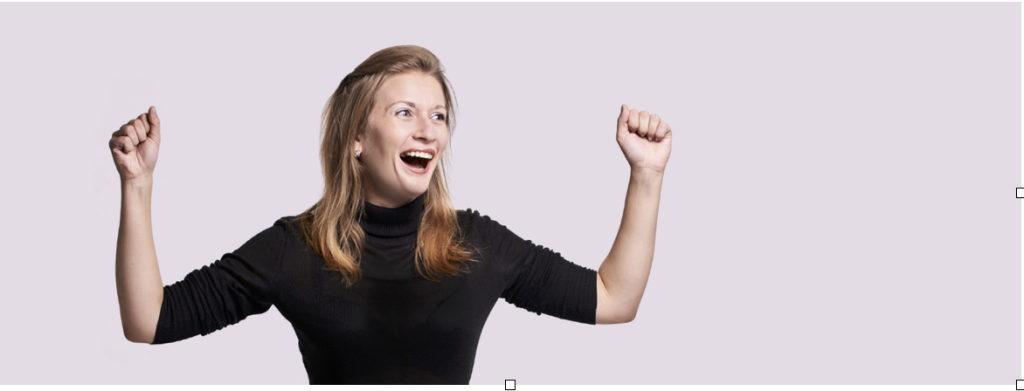 A woman with light brown hair, wearing a black turtleneck, is standing with her arms raised and a joyful, excited expression on her face. The background is a soft pastel purple color.